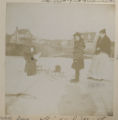 Judith Hartley standing in the snow holding a doll with Irma Hartley on a pair of skis and Ida Nelson watching