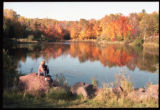 A person sitting on a rock overlooking a pond at Bagley Nature Center in fall